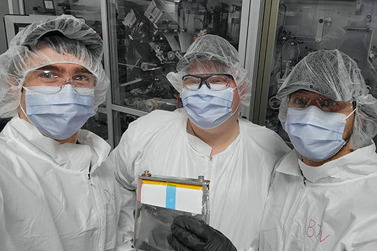 Three researchers wear face masks and plastic protective hair and body covers in a lab.