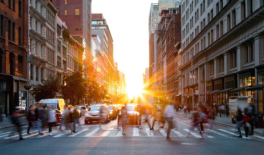 With the sun setting, pedestrians cross a sidewalk in an urban setting with waiting cars in the background.