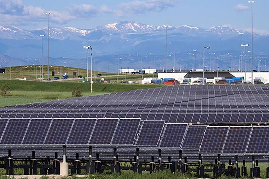 Solar panels in a field with buildings and mountains in the background.