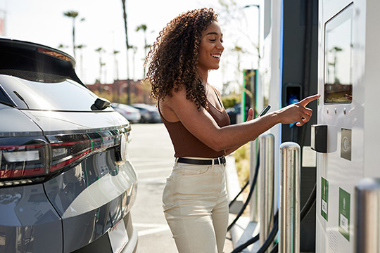 A person reaches for a button at an EV charging station; a car is plugged in behind her.