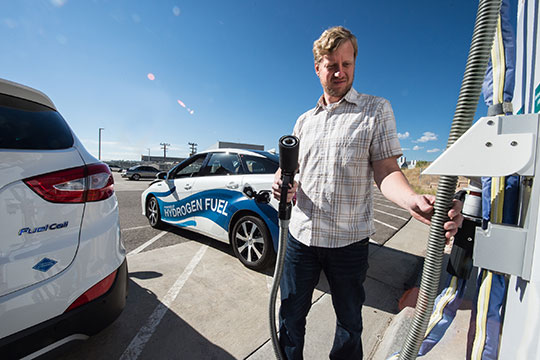 A man stands in front of a hydrogen fuel cell car as he begins to fuel it with a nearby hydrogen station.
