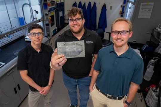 Three researchers smile for a photo in a lab. The person in the center holds a small carbon fiber composite sample with a black-and-white woven design and a raised indent with multiple sides.