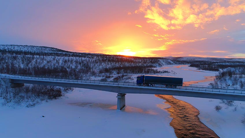 Semi-truck crosses bridge over icy river with sunset in the background.
