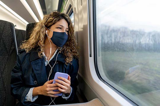 A person sitting on a train wearing headphones and a mask.
