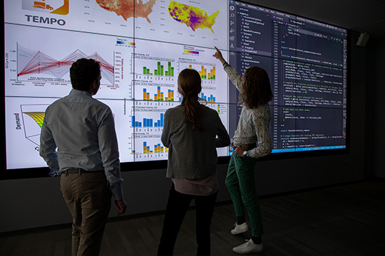 Three researchers with their backs to the camera look at a large screen with charts and graphs on it.