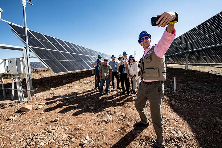 A man in a hardhat takes a selfie with a group of individuals behind him. They are all standing on dirt in a field of PV panels.