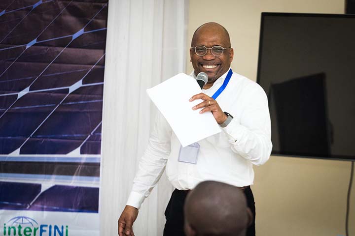 Man holding microphone and piece of paper while smiling and talking to an audience at the Greening the Grid Tanzania workshop. A screen is behind him.