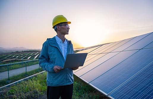 A man in a hard hat holding a computer and standing in a solar farm.