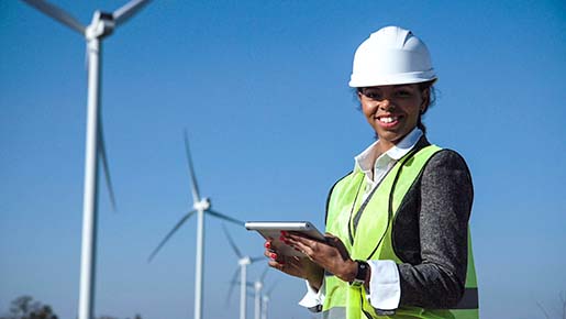 A woman in a hardhat and reflective vest holds a tablet and stands in front of wind turbines.