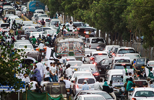 On a crowded Pakistan street, cars are in traffic with many people around