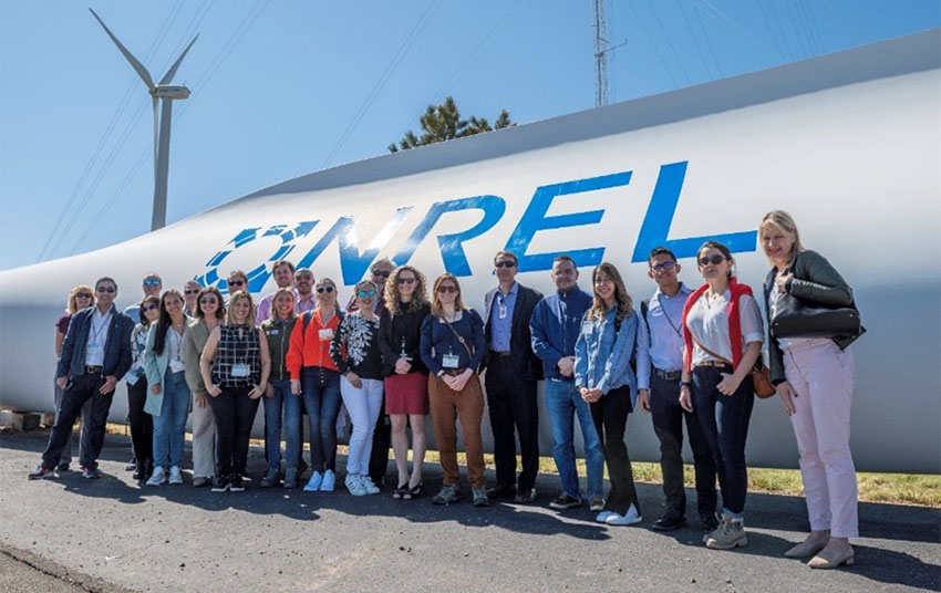 Add photo of group in front of turbine. alt="Training Program participants smile in front of a wind turbine blade marked with the NREL logo.