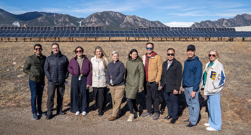 Add photo: alt="Study tour visitors smile for the camera in front of a solar array and the Flatirons