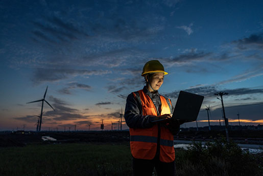 A woman with personal protective equipment smiles at her laptop in a wind farm at dusk
