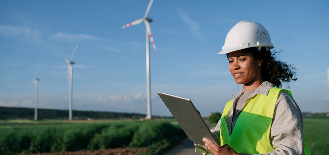 Person wearing hardhat, stands in field examining clipboard with wind turbines in the background.