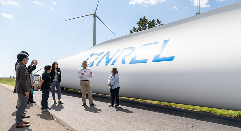 Tour group listens to a guide in front of a wind turbine blade branded with the NREL logo.