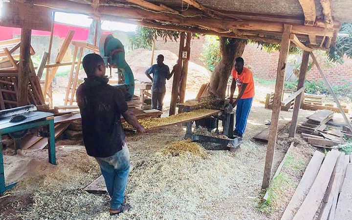 Men running a wooden board through a band saw under thatched-roof.