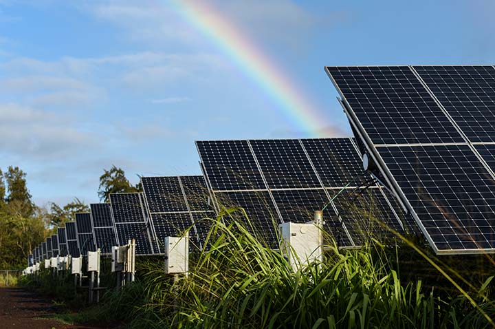 A ground mounted solar array in a green field, with a blue sky and rainbow behind it.