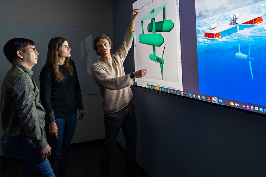 Three researchers look at marine energy prototypes on a large display.