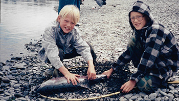 Two kids hold fish in net on shoreline.
