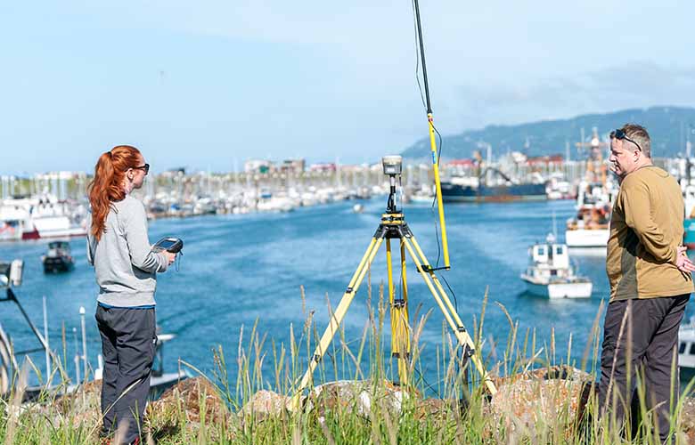Two people collecting data from a marine energy device on a cliff above a harbor.
