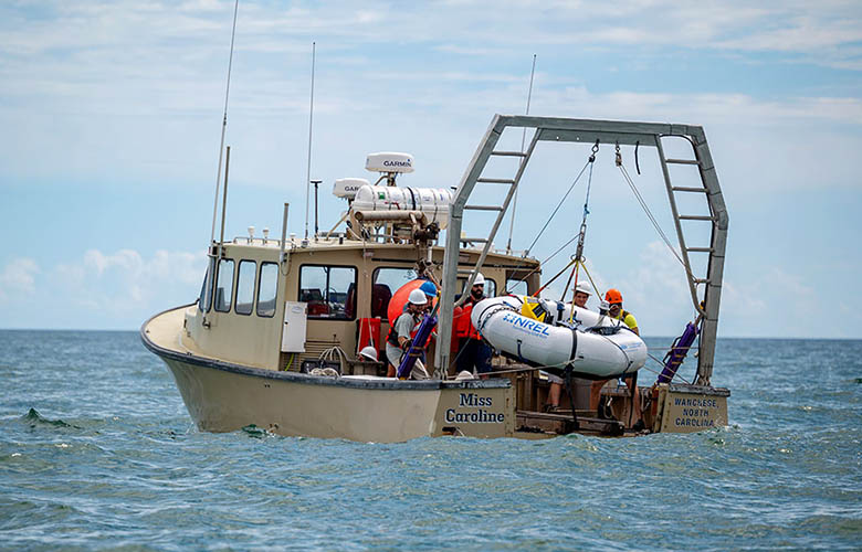 An inflatable hexagonal tube being lowered from a crane on the back of a boat into the ocean.