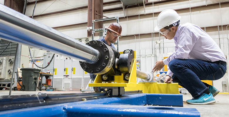 Two people in hard hats work on a large mechanical device on lab floor.
