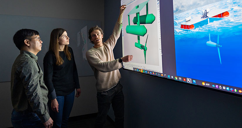 Three researchers looking at an enlarged computer screen featuring illustrations of two tidal energy devices.