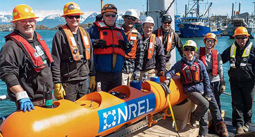 A group of people in hard hats and life vests stand on a dock next to a buoy painted with NREL’s logo.
