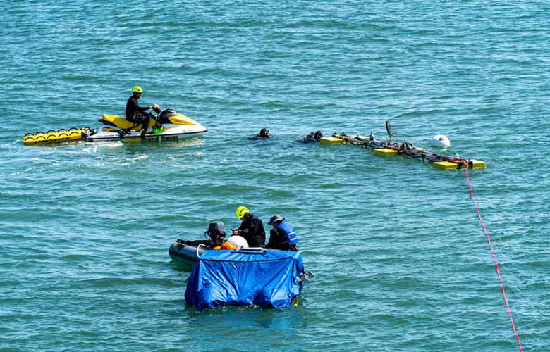 Two scuba divers secure a wave energy device to its anchor site as people watch from a jet ski and raft.