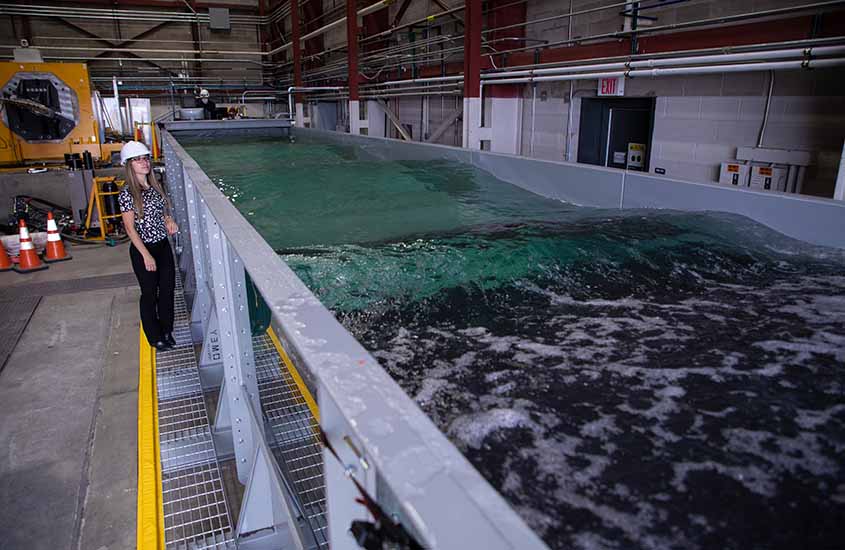 A woman in hard hat stands next to an indoor wave tank.
