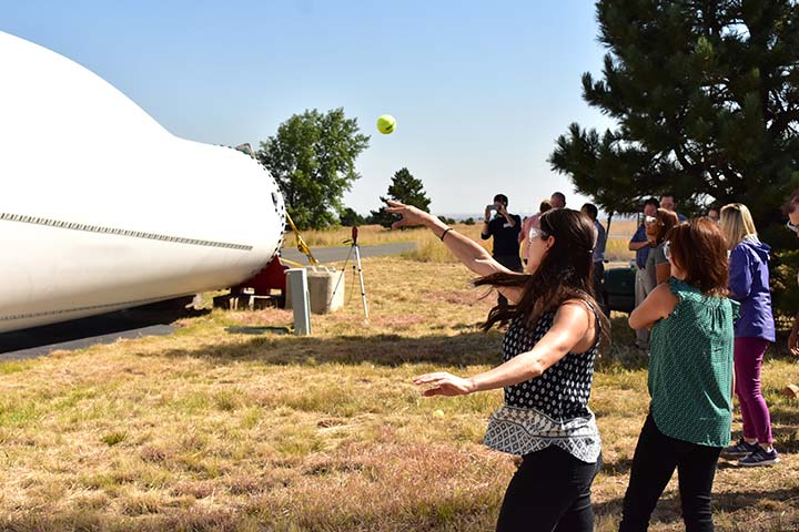 A woman throws a tennis ball at turbine blade with attached sensor equipment on the ground.