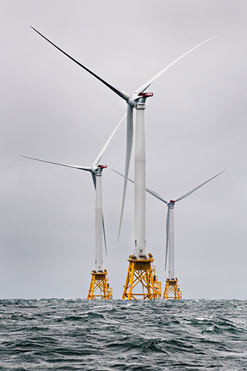 A group of wind turbines stand over ocean waves.