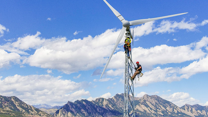 Two people climb a wind turbine with mountains in the background.