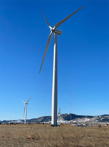 A tall turbine stands in a field; two smaller turbines and hills are in the background.