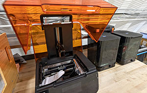 An orange translucent cover is lifted off a black, rectangular device that sits on a desktop, revealing some wires and moving parts inside.