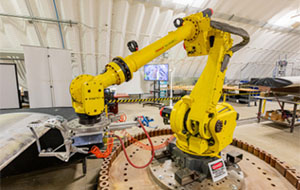 A large, yellow robotic arm sits on a rotating dias in the middle of a hangar.