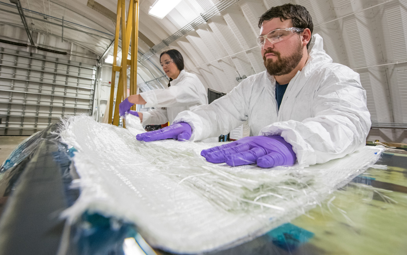 Two people wearing safety goggles, lab coats, and gloves placing composite materials in a turbine blade mold.