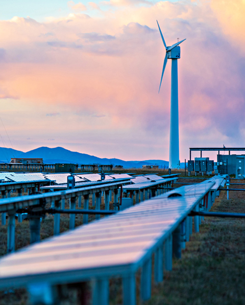 A wind turbine behind a group of solar panels during sunset.