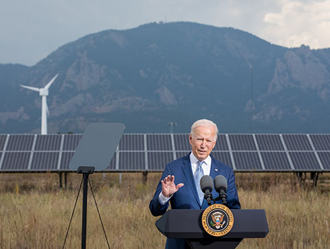 President Biden stands behind a podium in front of solar panels, a wind turbine, and mountains.