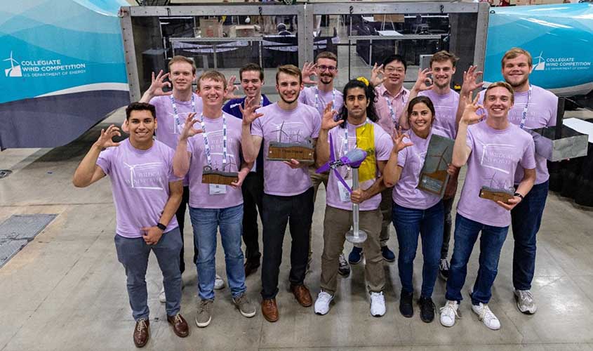 Students in matching shirts with a matching hand gesture holding a model wind turbine in front of a wind tunnel and Collegiate Wind Competition signs.