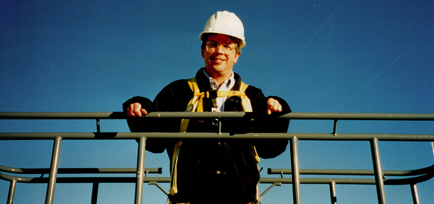 A man stands against a railing wearing a hard hat.