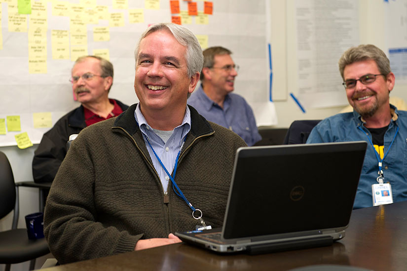A man smiles as he sits in front of an open laptop. Three men sit smiling behind him.
