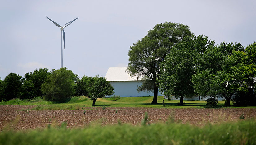 A small wind turbine towers over a field, forest, and barn on a farm.