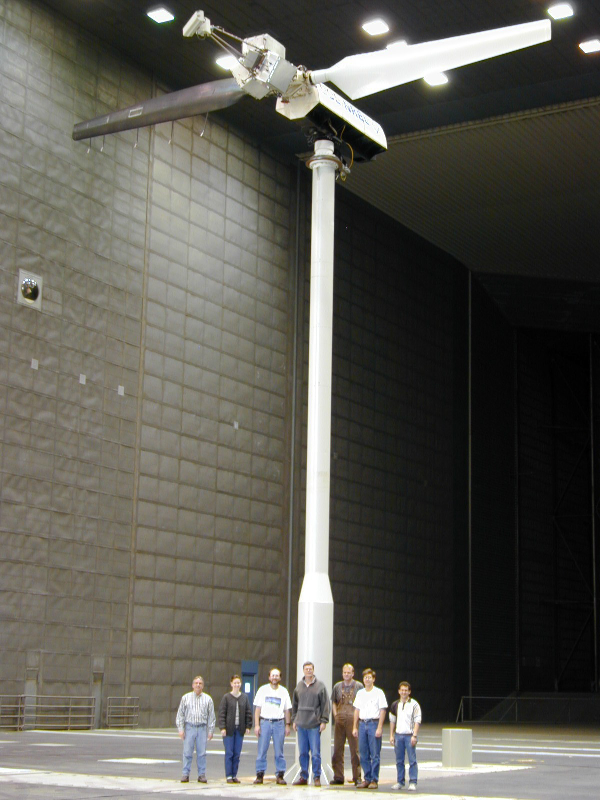 A two-bladed wind turbine inside a large garage-like space with seven people standing in the foreground.