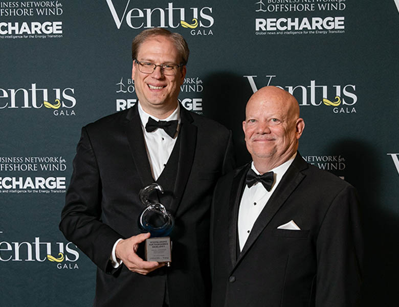 Two people in formal suits, one holding an award, stand in front of a backdrop with the logos of the Ventus Gala, Business Network Offshore Wind, and Recharge.