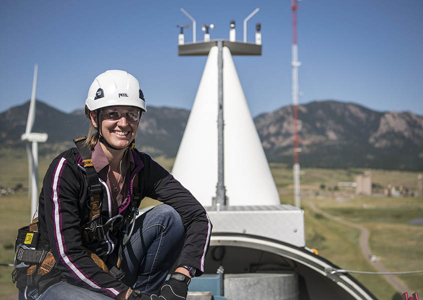 Jen crouches on the top of a wind turbine in a hard hat, goggles, and safety harness.