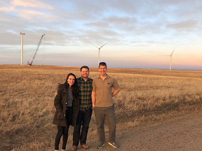 Three people stand in front of wind turbines in a field.