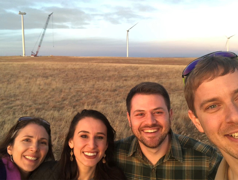 Four people smiling with wind turbines behind them in a field.