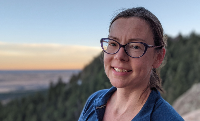 Julie Lundquist in glasses and a ponytail at the top of a mountain with blurry trees and sunset in the background.