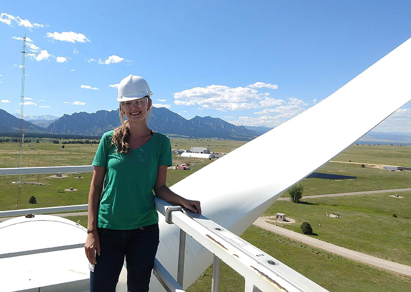 Jen in hard hat and goggles stands in front of a wind turbine blade above a campus. Mountains are in the distance.